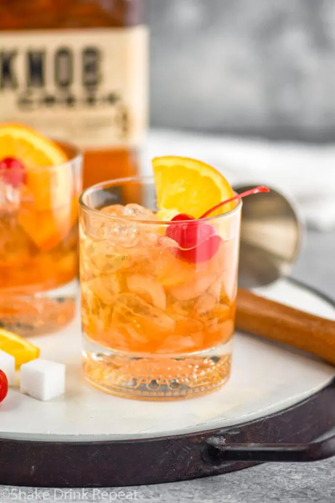 Two glasses of old fashioned with ice, cherry, orange slice and muddler sitting off to the side