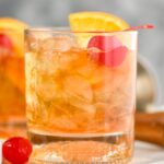 Glass of old fashioned with ice, cherry and orange on a tray with cherry and muddler