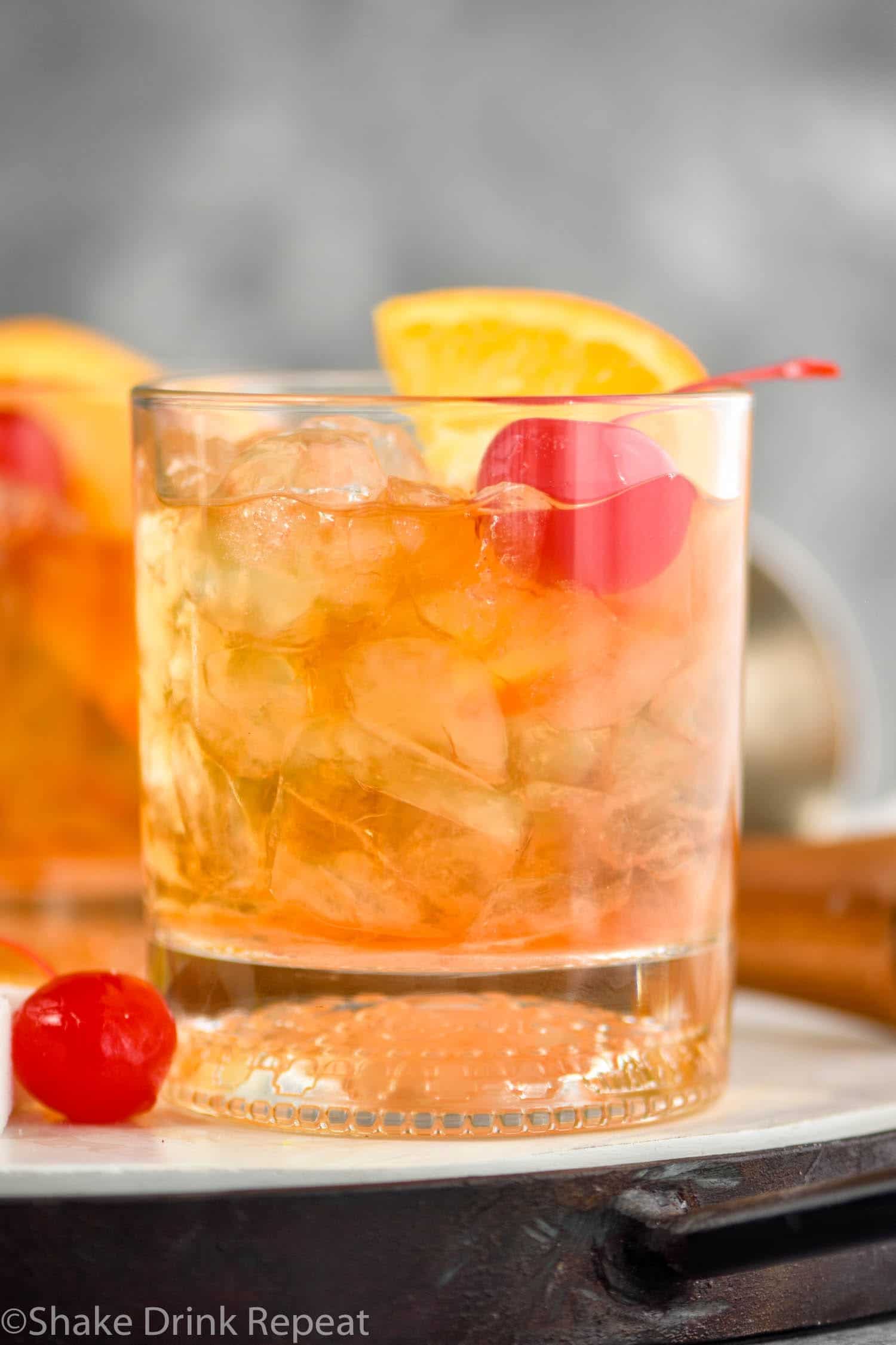 Glass of old fashioned with ice, cherry and orange on a tray with cherry and muddler