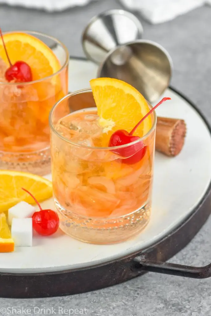 Two glasses of old fashioned with ice, cherry, orange slice and muddler sitting off to the side with a sugar cube