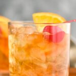 Two glasses of old fashioned with ice, cherry, orange slice and muddler sitting off to the side with a cherry