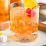 Two glasses of old fashioned with ice, cherry, orange slice and muddler sitting off to the side with a sugar cube