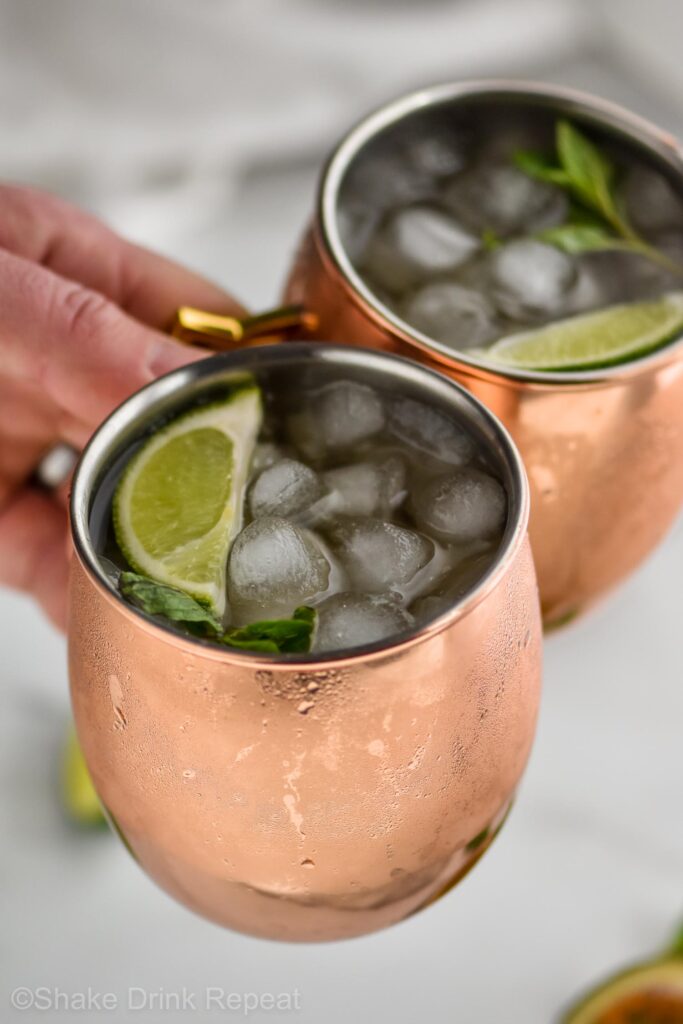 man holding two copper mugs of passion fruit moscow mule with ice, limes, and fresh mint leaves