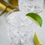 Two glasses of Vodka Press with ice, lime wedges, jigger and spoon off to the side