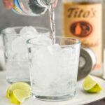 Two glasses of Vodka Soda with lime wedges and bottle of Tito's on the sides, bottle of club soda being poured