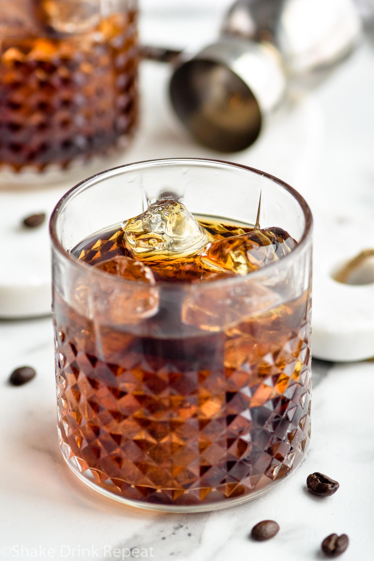 Two glasses of Black Russian with ice, surrounded by coffee beans and a jigger off to the side