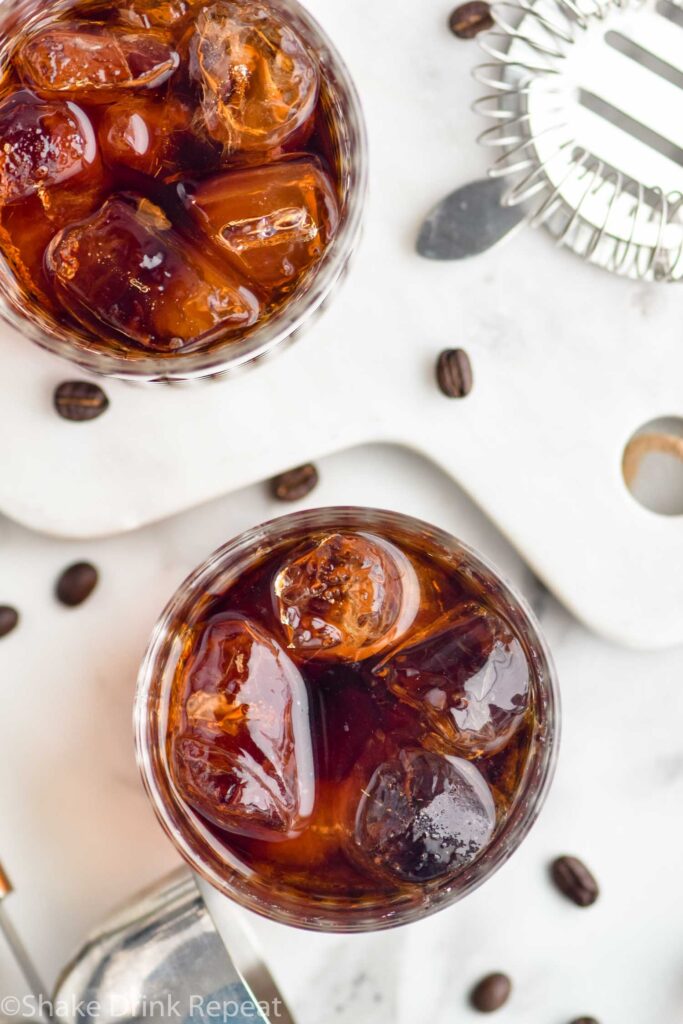 Two glasses of Black Russian with ice, surrounded by coffee beans and a strainer off to the side