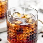 Two glasses of Black Russian with ice, surrounded by coffee beans and a strainer off to the side