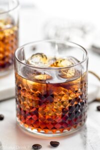 Two glasses of Black Russian with ice, surrounded by coffee beans and a strainer off to the side
