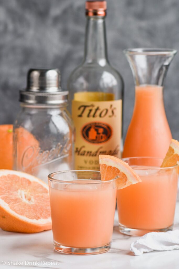 Two glasses of Greyhound Cocktail with ice and grapefruit wedge on the rim, bottle of Tito's vodka and shaker in background