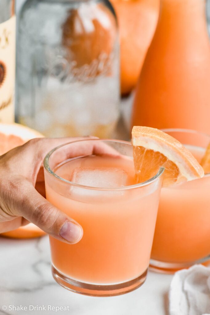 A glass of Greyhound Cocktail with a grapefruit wedge and ice cube with a pitcher in the background