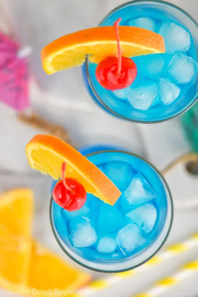 two glasses of blue lagoon cocktail with ice, orange slice and cherry garnish