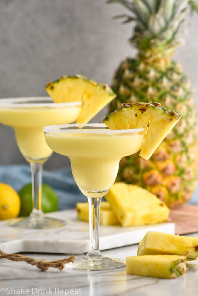 two glasses of pineapple margarita with salted rim surrounded by lemons and limes