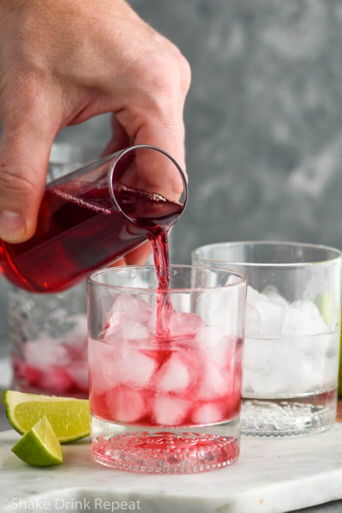 hand pouring cranberry juice into glass of vodka cranberry recipe with ingredients in background