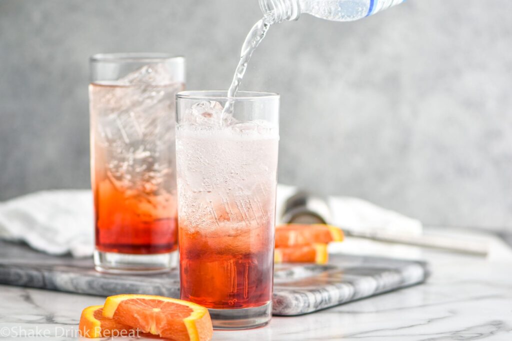 two glasses of americano cocktail ingredients with ice, orange slices and club soda splash