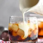 Glass of white russian cocktail with ice and cream being poured surrounded by coffee beans