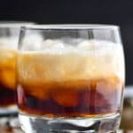 Glass of white russian cocktail with ice surrounded by coffee beans