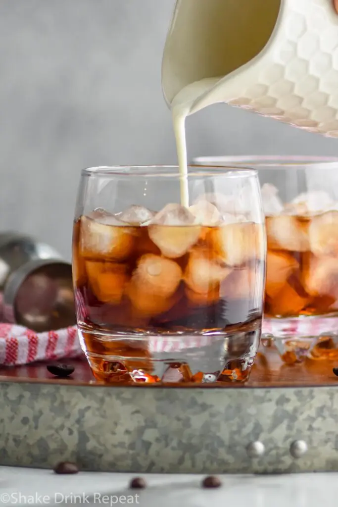 Glass of white russian cocktail with ice and cream being poured surrounded by coffee beans