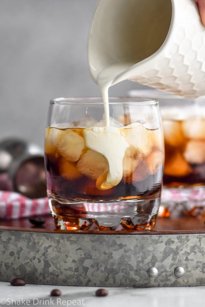 Glass of white russian cocktail with ice and cream being poured surrounded by coffee beans