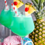 two glasses of blue hawaii with ice, straws, pineapple, and cherry