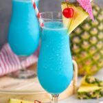two glasses of frozen blue hawaiian with straw, umbrella, pineapple, and cherry garnish