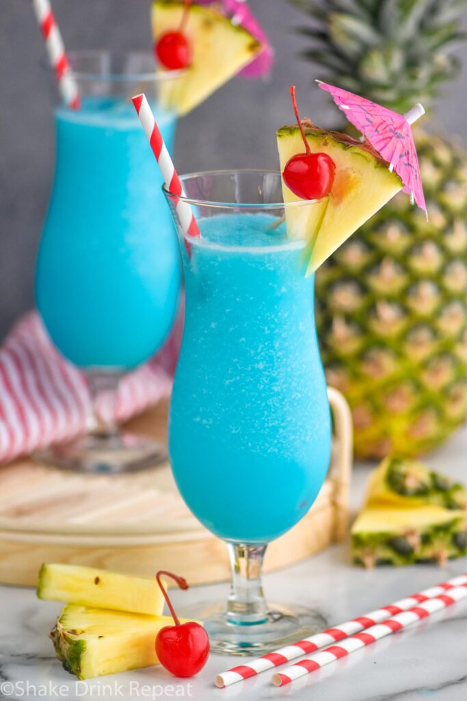 two glasses of frozen blue hawaiian with straw, umbrella, pineapple, and cherry garnish