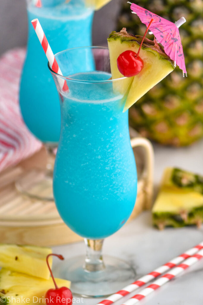 two glasses of frozen blue hawaiian with straw, umbrella, pineapple, and cherry