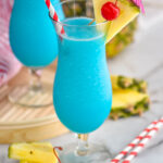 two glasses of frozen blue hawaiian with straw, umbrella, pineapple, and cherry garnish