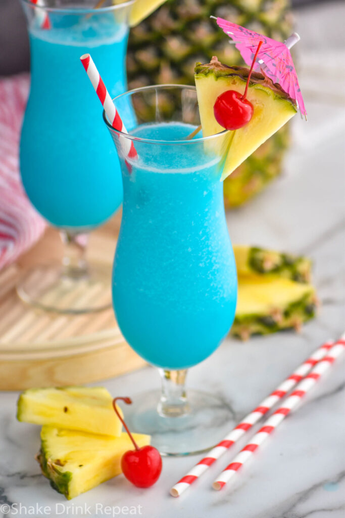 two glasses of frozen blue hawaiian with straw, umbrella, pineapple, and cherry garnish