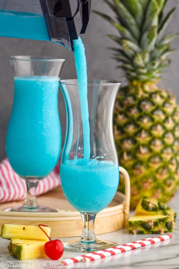 pouring a frozen blue hawaiian drink from a blender into a glass with pineapple and cherry garnish