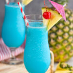 two glasses of frozen blue hawaiian with straw, umbrella, pineapple, and cherry garnish