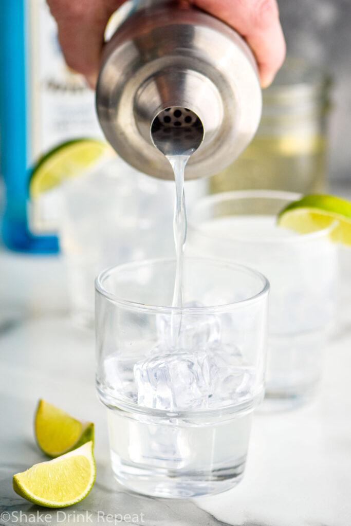 man pouring gin gimlet recipe from a shaker into a glass filled with ice and surrounded by lime wedges