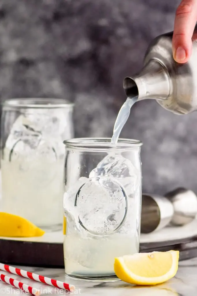making two glasses of long island iced tea by pouring ingredients from shaker over ice with lemon garnish