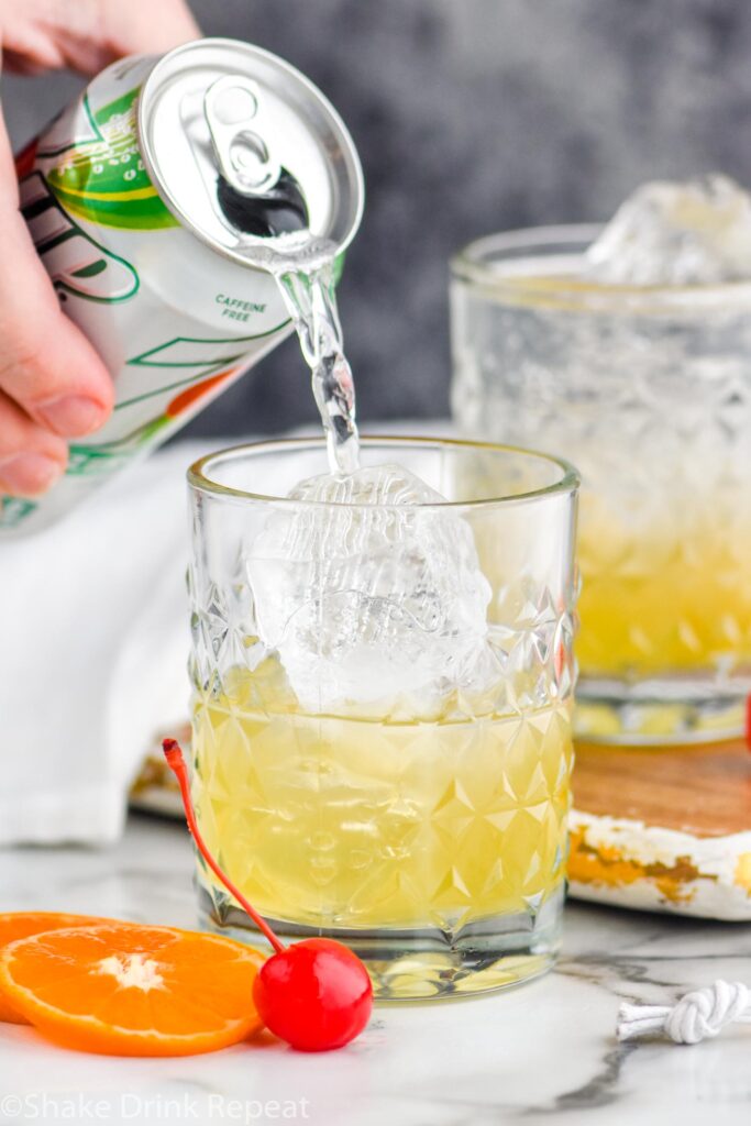 Man pouring lemon lime soda into a glass of amaretto sour ingredients with ice, surrounded by orange slices and a cherry