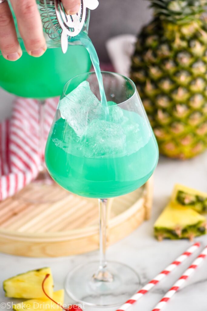 pouring a blue hawaii into a cocktail glass with ice, pineapple and cherry garnish