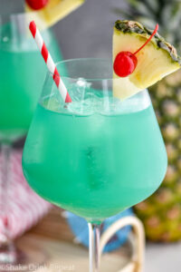 glass of blue hawaii with ice, a straw, pineapple, and a cherry