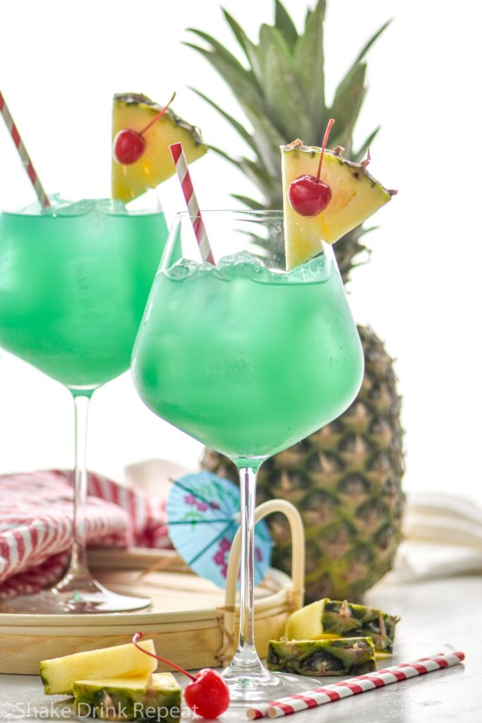 two glasses of blue hawaii with ice, straws, pineapple, and cherry garnish
