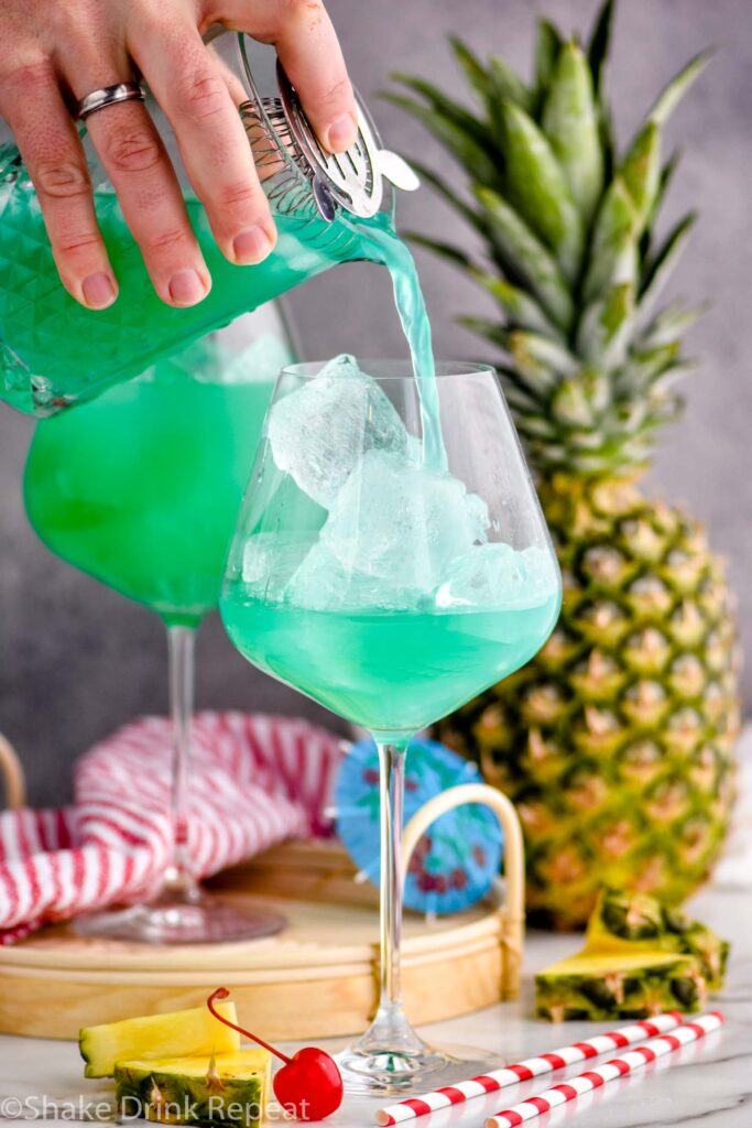 pouring a blue hawaii into a cocktail glass with ice surrounded by pineapple and a cherry.