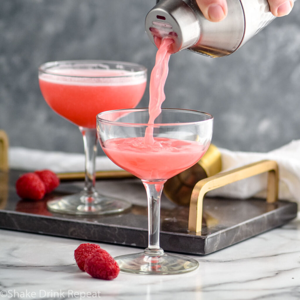 man pouring clover club cocktail from a shaker into a cocktail glass with raspberries