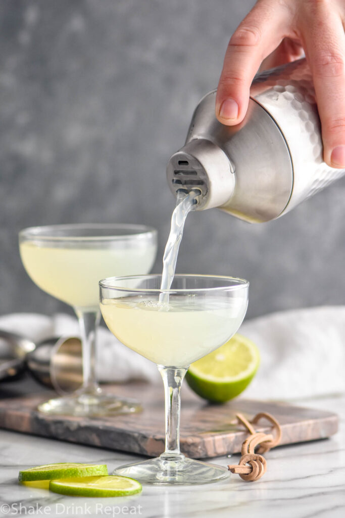 man pouring daiquiri from a shaker into a chilled cocktail glass surrounded by limes