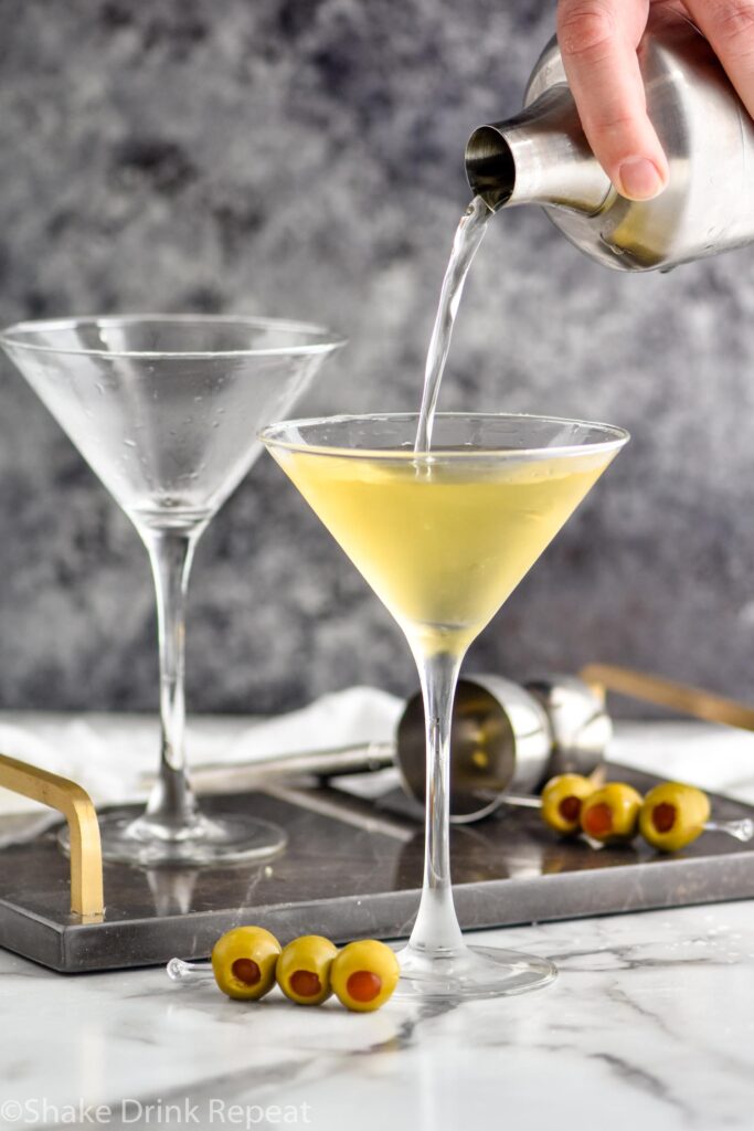 pouring a dirty martini into a martini glass with green olives