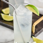 glass of gin rickey with ice and straw surrounded by lime wedges