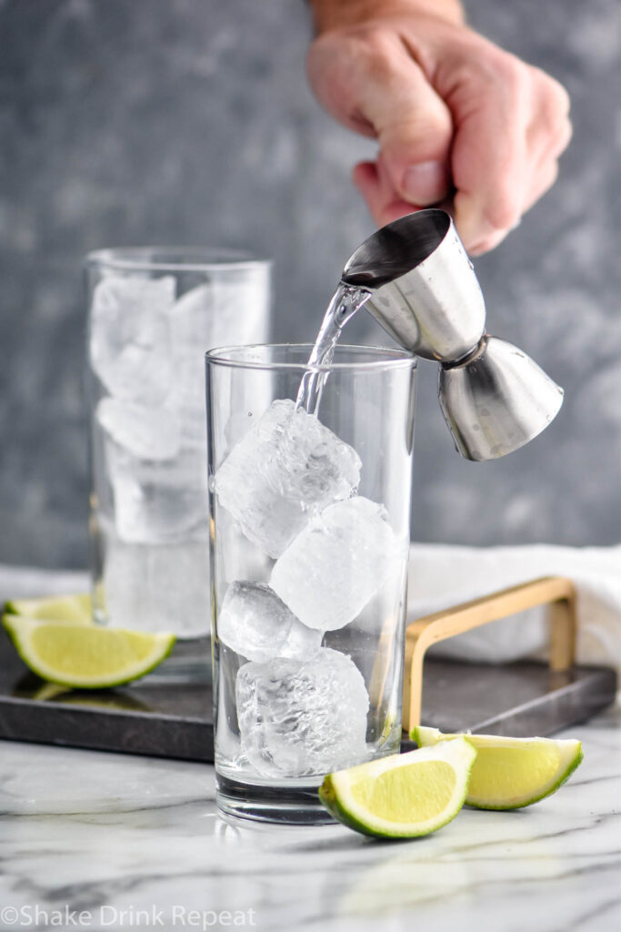 man pouring jigger of gin into a glass of ice to make a Gin Rickey surrounded by lime wedges