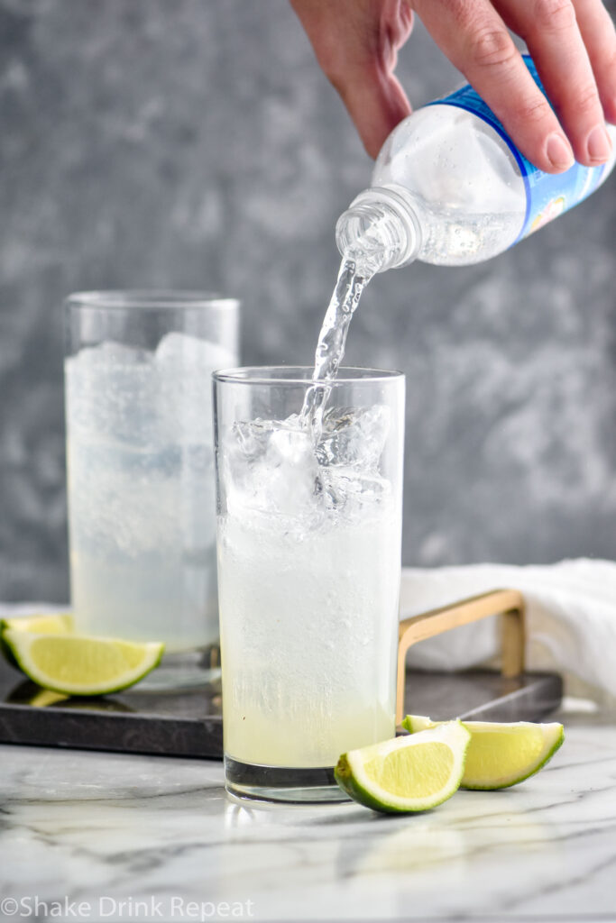 man pouring club soda water into a glass of Gin Rickey, ice, and lime wedges.