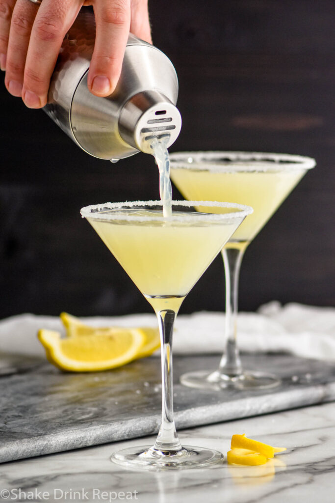 man's hand pouring lemon drop martini into a martini glass with sugared rim and lemon twist garnish