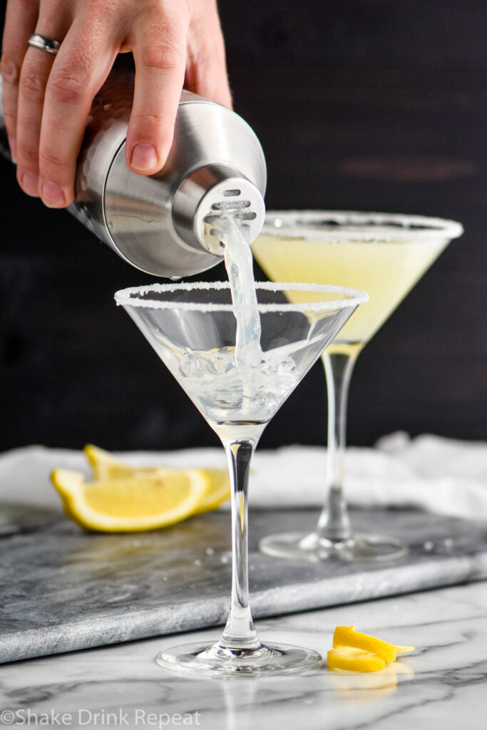 man pouring a lemon drop martini into a glass rimmed with sugar surrounded by lemon slices