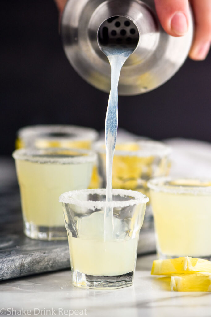 man pouring lemon drop shot from a shaker into shot glasses with sugared rim and lemon wedge