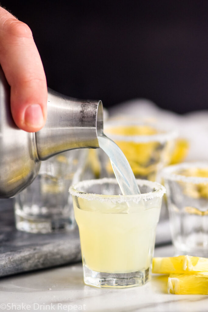man pouring lemon drop shot from a shaker into a shot glass with sugared rim and lemon wedges