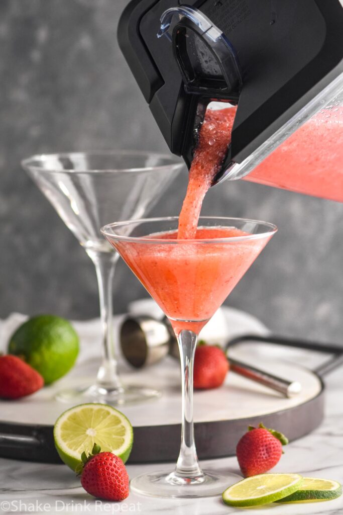 pouring strawberry daiquiri from a blender into a cocktail glass with fresh limes and strawberries