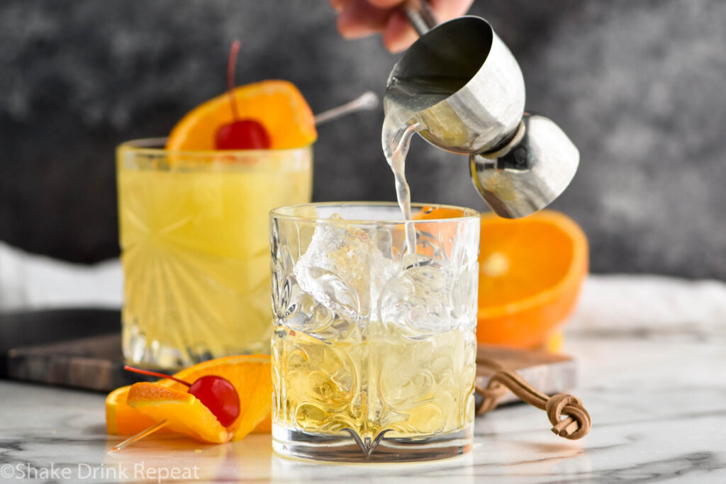 jigger pouring ingredients into of Amaretto Stone Sour into a glass filled with ice surrounded by orange slice and cherry garnish